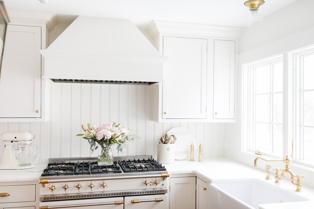 Bright white kitchen with a large stove, gold accents, and a well-organized Kitchen Counter Organization featuring a bouquet of flowers. Plenty of natural light pours through the windows, highlighting the tidy elegance.