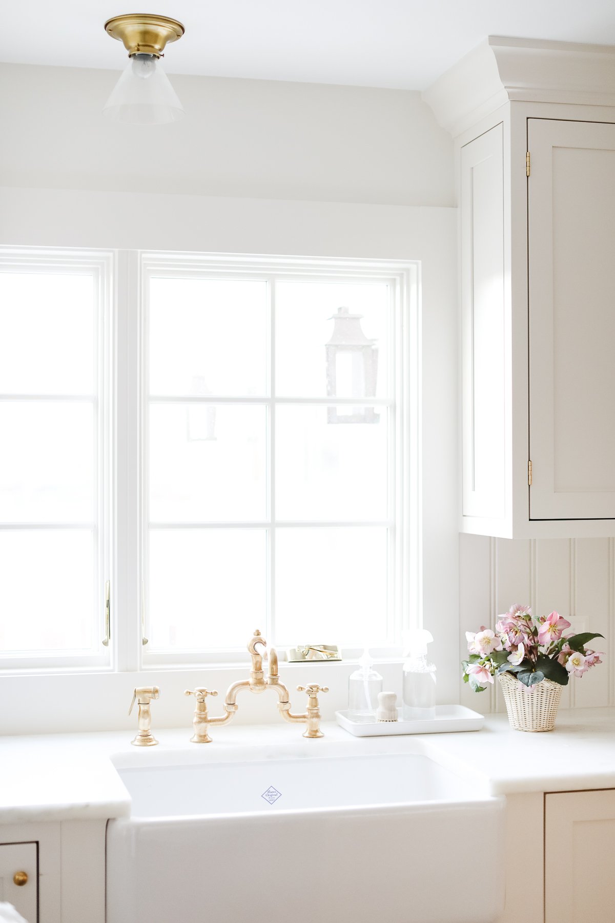 A bright kitchen features a white farmhouse sink, brass faucet, and pristine cabinets. The kitchen counter organization is complemented by a vase of pink flowers near the large window, adding a touch of charm.