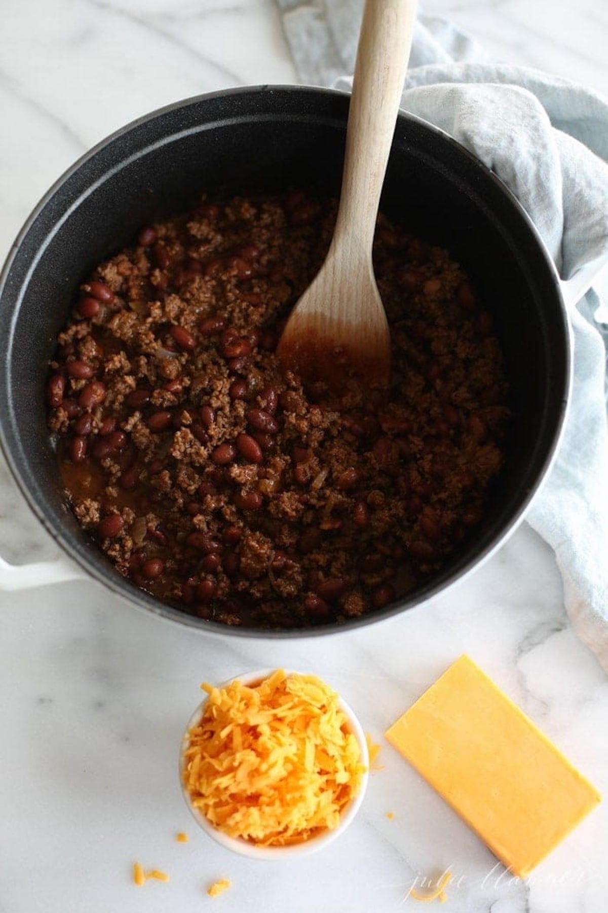 A quick and easy chili recipe cooked in a pan, topped with melted cheese and stirred with a wooden spoon.