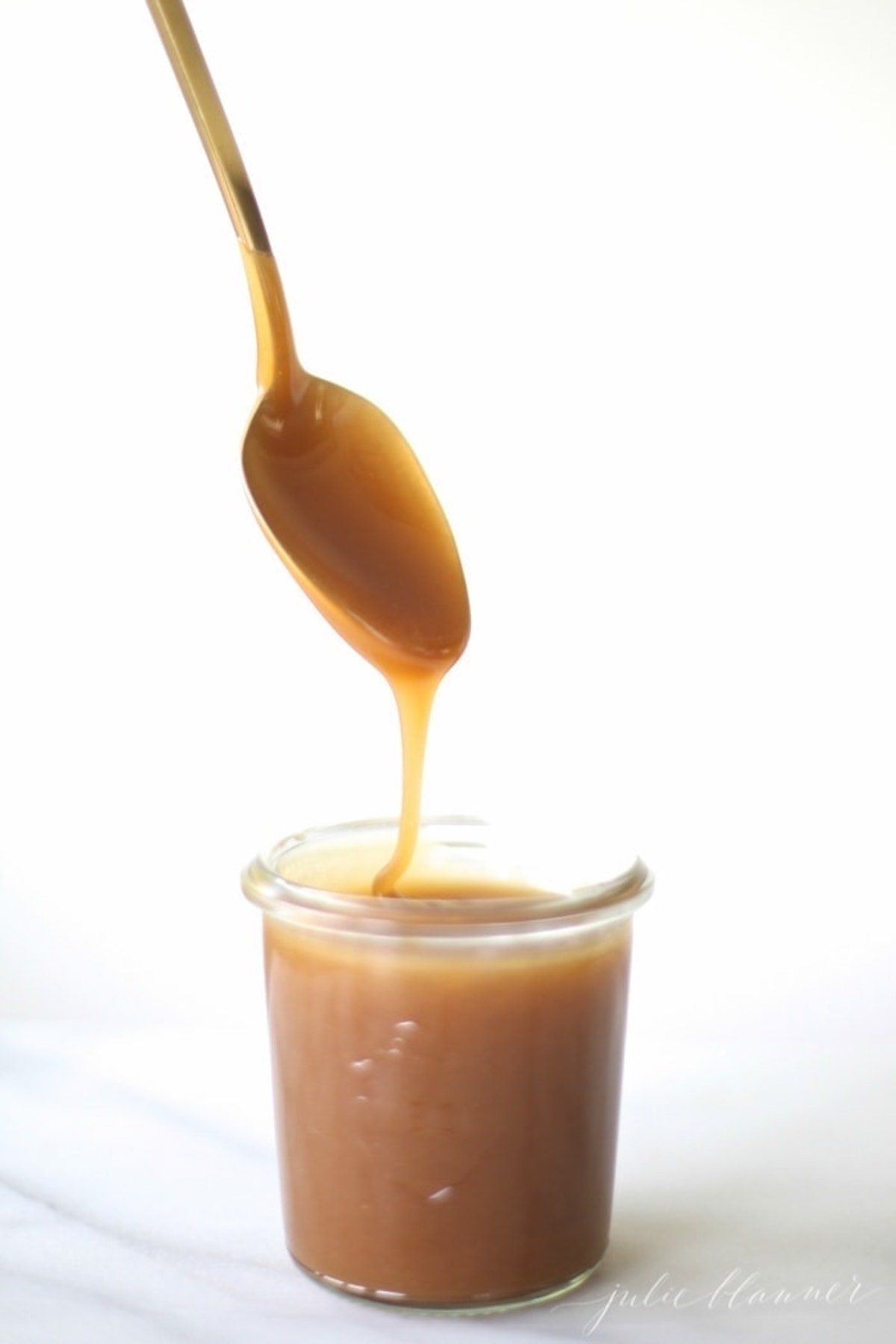 A spoon is pouring caramel sauce, specifically salted caramel sauce, into a jar.