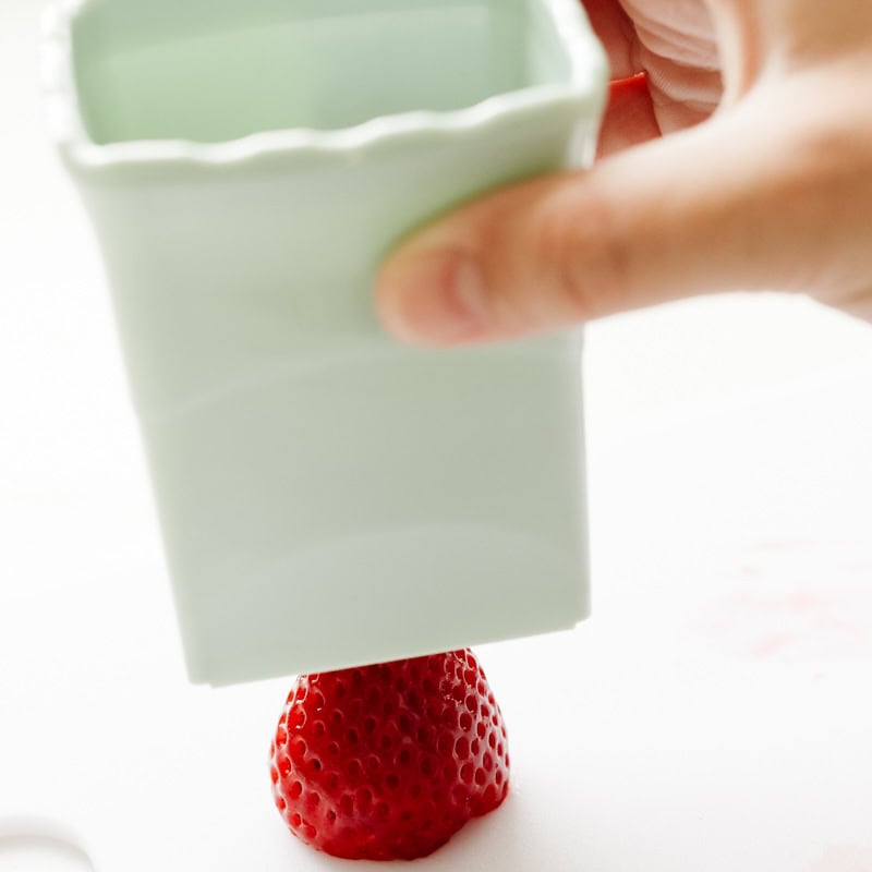 A hand utilizes an innovative kitchen gadget, pressing a rectangular white container over a strawberry on a pristine white surface.