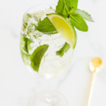 A glass of Hugo spritz with mint leaves and a lime wedge, accompanied by a gold-colored stirring spoon on a marble surface.