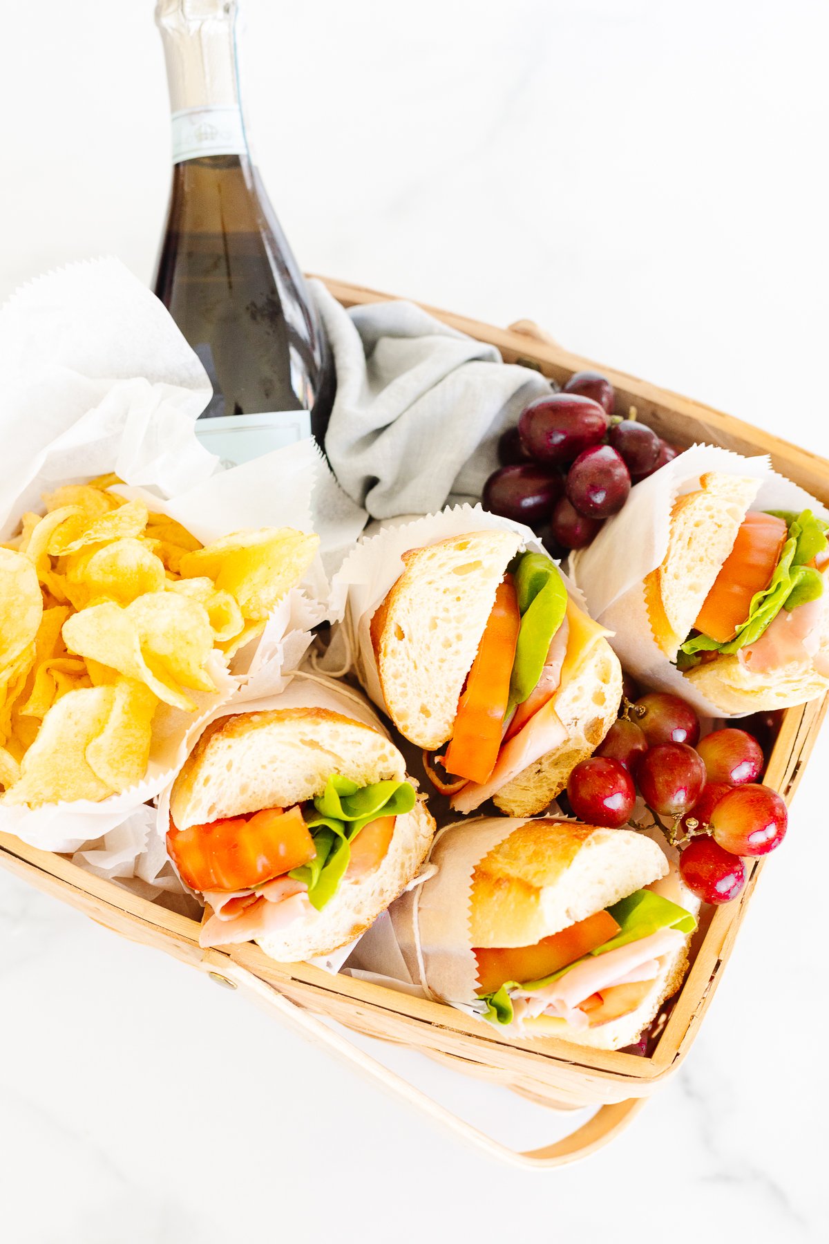 A picnic basket containing sandwiches, chips, grapes, and a bottle of sparkling beverage.