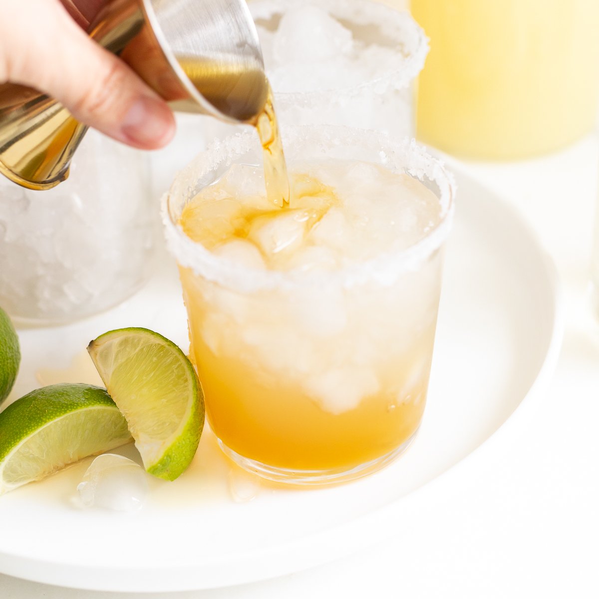 Pouring a Cadillac margarita into a salt-rimmed glass with lime wedges and a pitcher in the background.