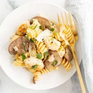 A bowl of chicken and mushroom pasta, garnished with herbs, served with a fork on the side, on a white surface.