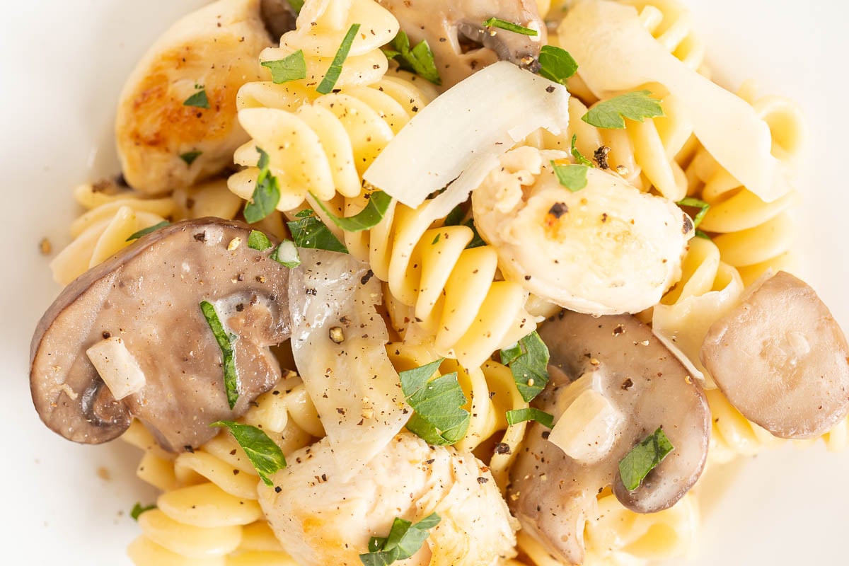 Close-up of a chicken mushroom pasta dish with grilled chicken, sliced mushrooms, and chopped herbs on a white plate.