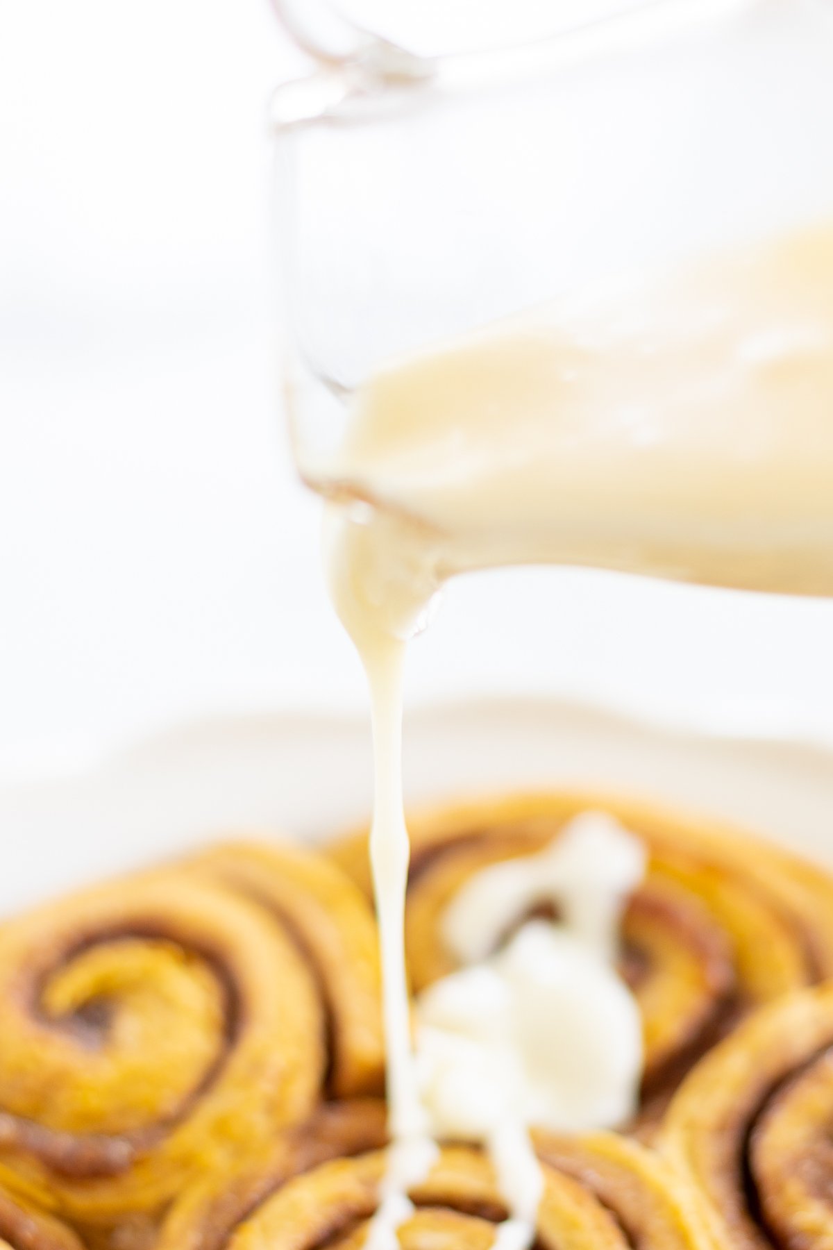 Cream cheese icing for cinnamon rolls pouring over a plate of pumpkin cinnamon rolls. 