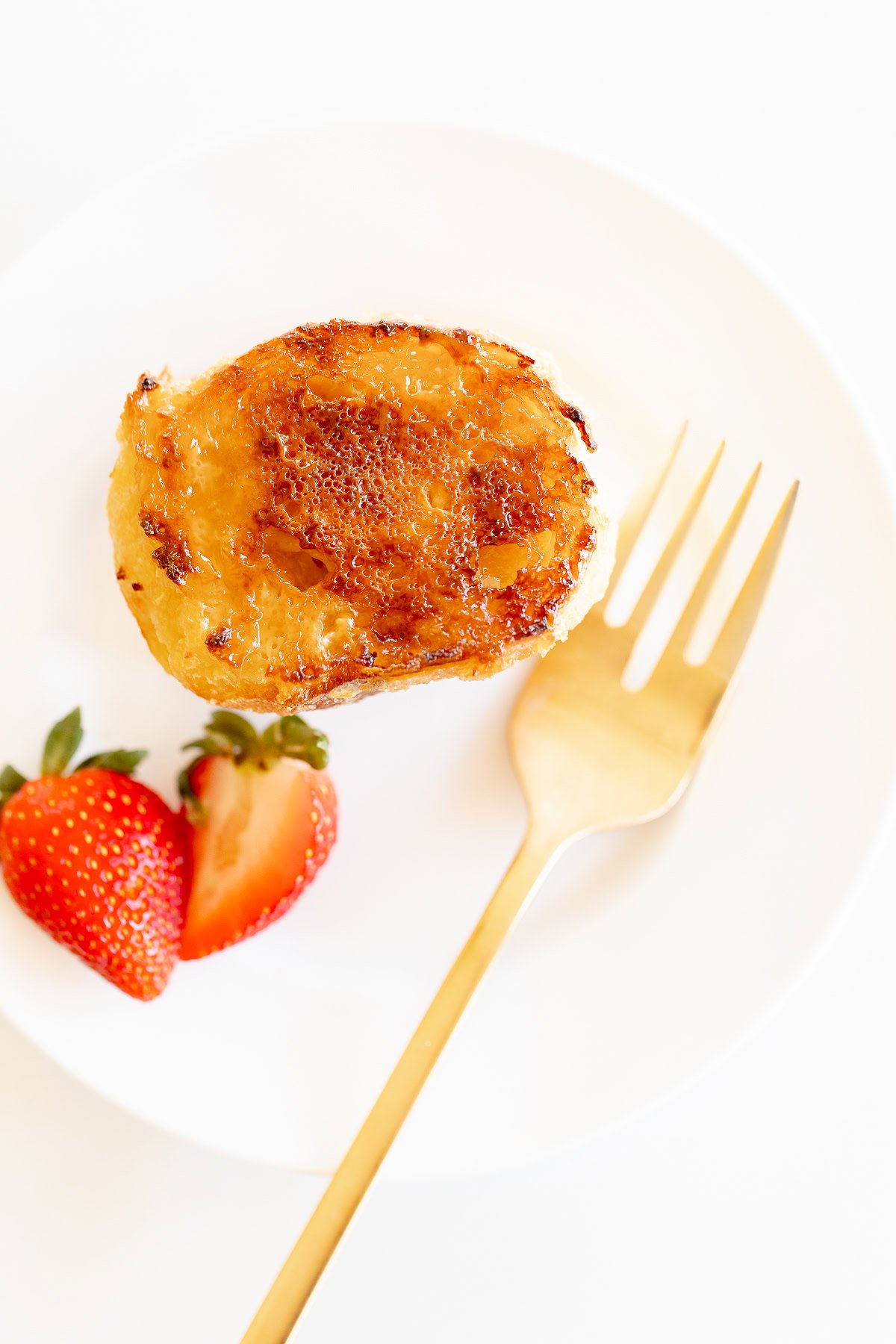 Creme Brulee French toast slice on a white plate with a gold fork. Garnished with sliced strawberries.