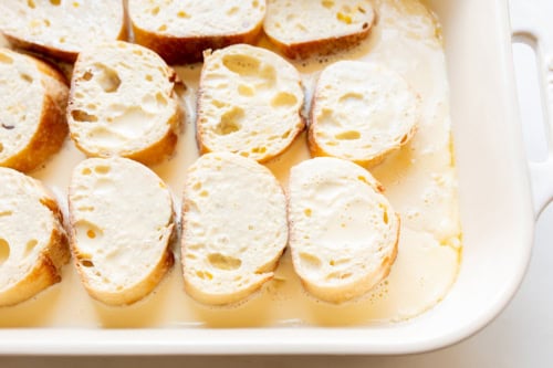 Slices of bread soaking in egg mixture in a white baking dish, preparing for Crème Brûlée French Toast Casserole.