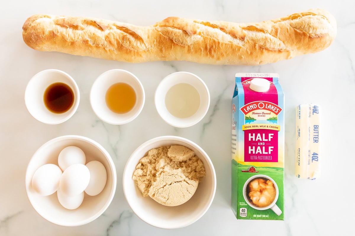 Ingredients for baking a Crème Brûlée French Toast Casserole displayed on a marble countertop, including eggs, flour, butter, half-and-half, and a baguette.