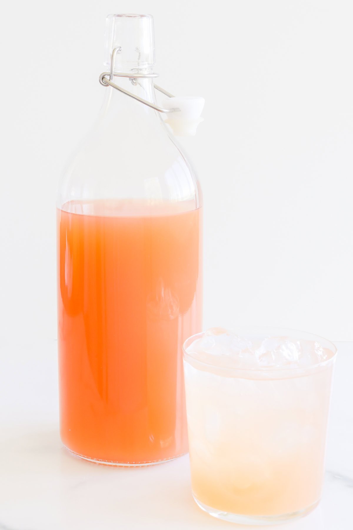 A clear glass bottle with a metal clasp containing paloma mix, next to a glass filled with a paloma cocktail.