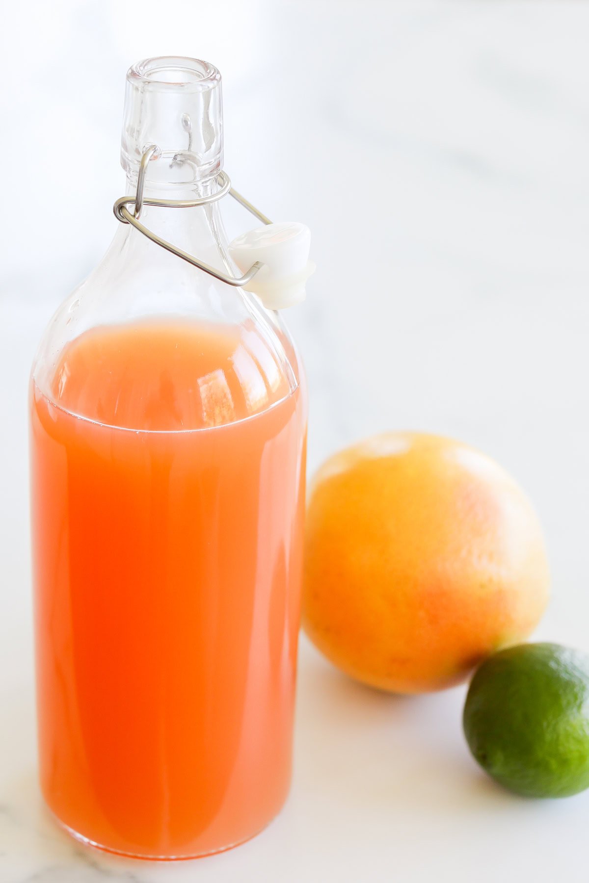 A glass bottle filled with orange juice, sealed with a ceramic stopper, next to a whole grapefruit and lime on a marble surface.