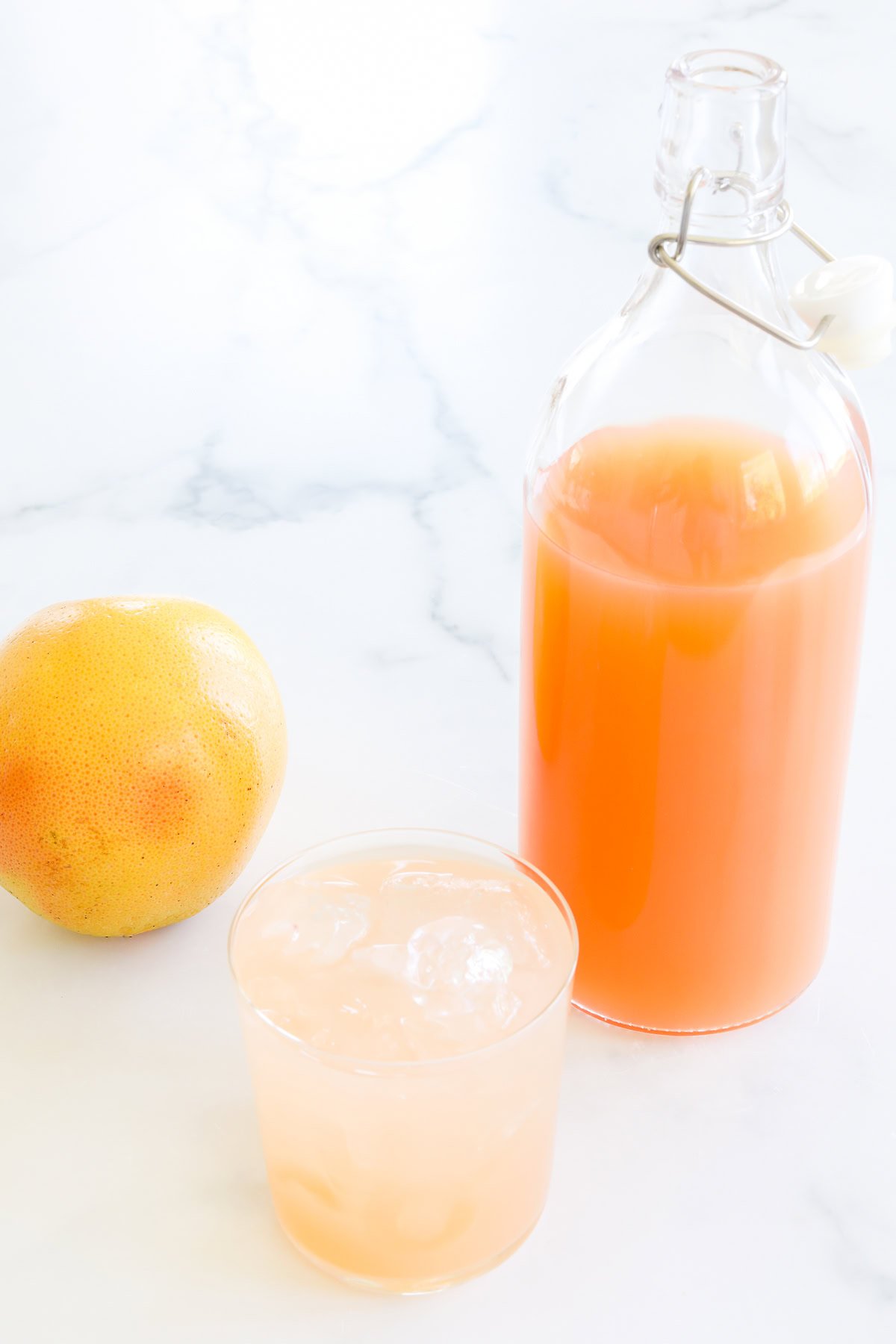 A glass bottle filled with paloma mix, next to a full glass with ice and a whole grapefruit on a marble surface.