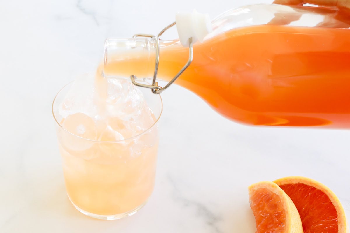 Paloma mix being poured from a glass bottle into a glass filled with ice cubes on a marble surface, next to sliced grapefruit.