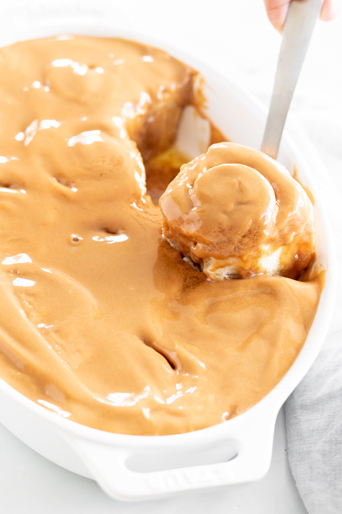 A hand serving a scoop of caramel-drizzled bread pudding from a white dish.
