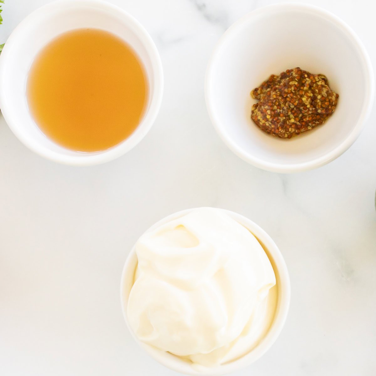 Three small bowls containing vinegar, whole grain mustard, and mayonnaise for a creamy pasta salad dressing recipe, on a marble surface.