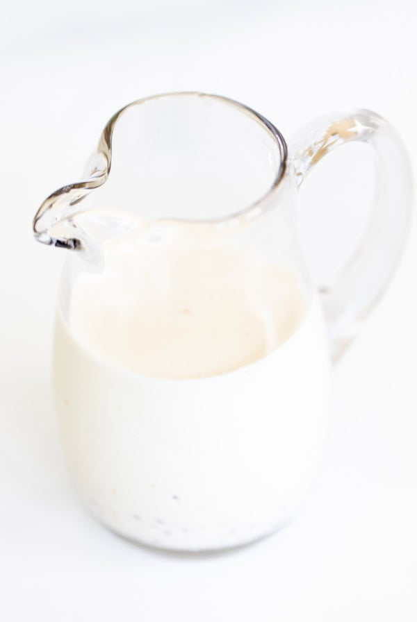 Clear glass pitcher filled with creamy pasta salad dressing on a white background.