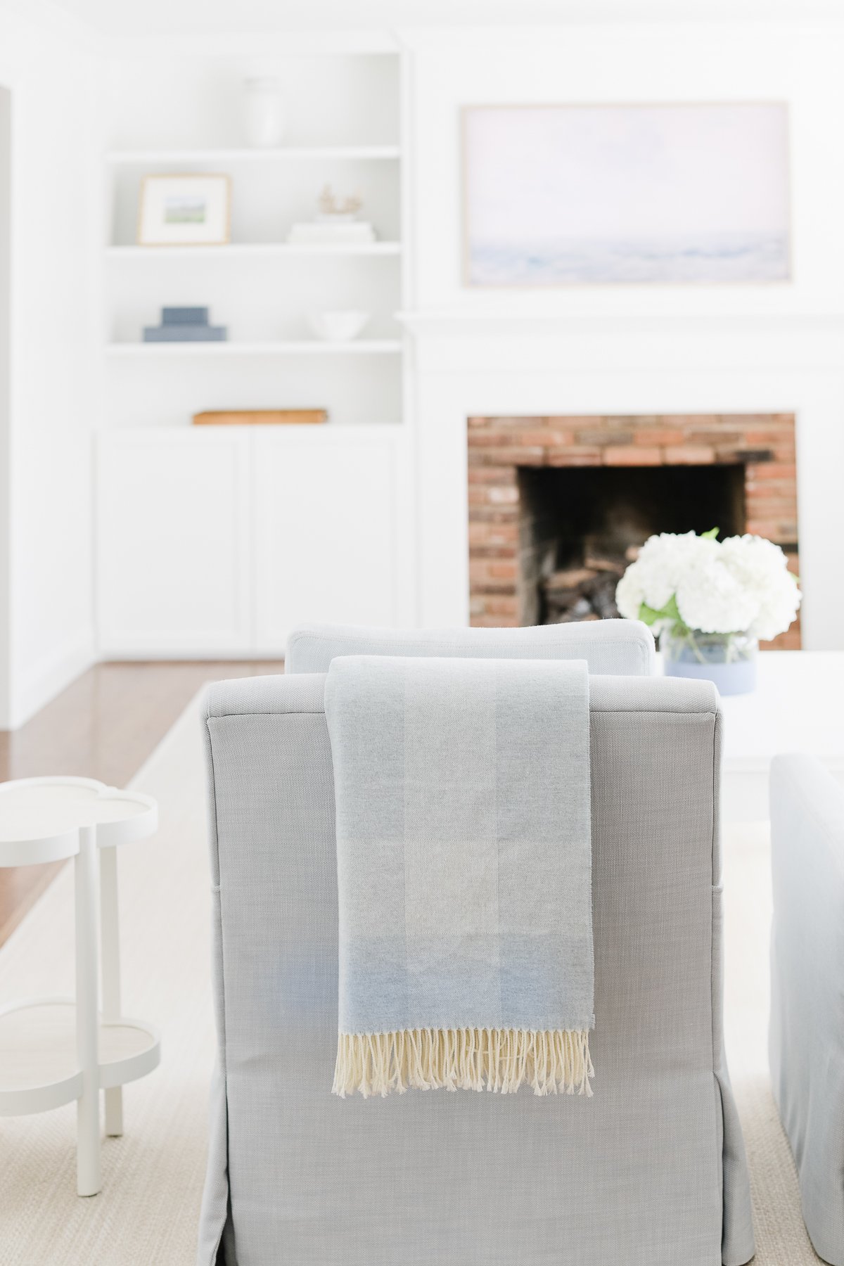 A cozy living room featuring a grey armchair with a blue throw draped over it, a brick fireplace, white bookshelves, and a light pink and blue framed artwork above the mantle.