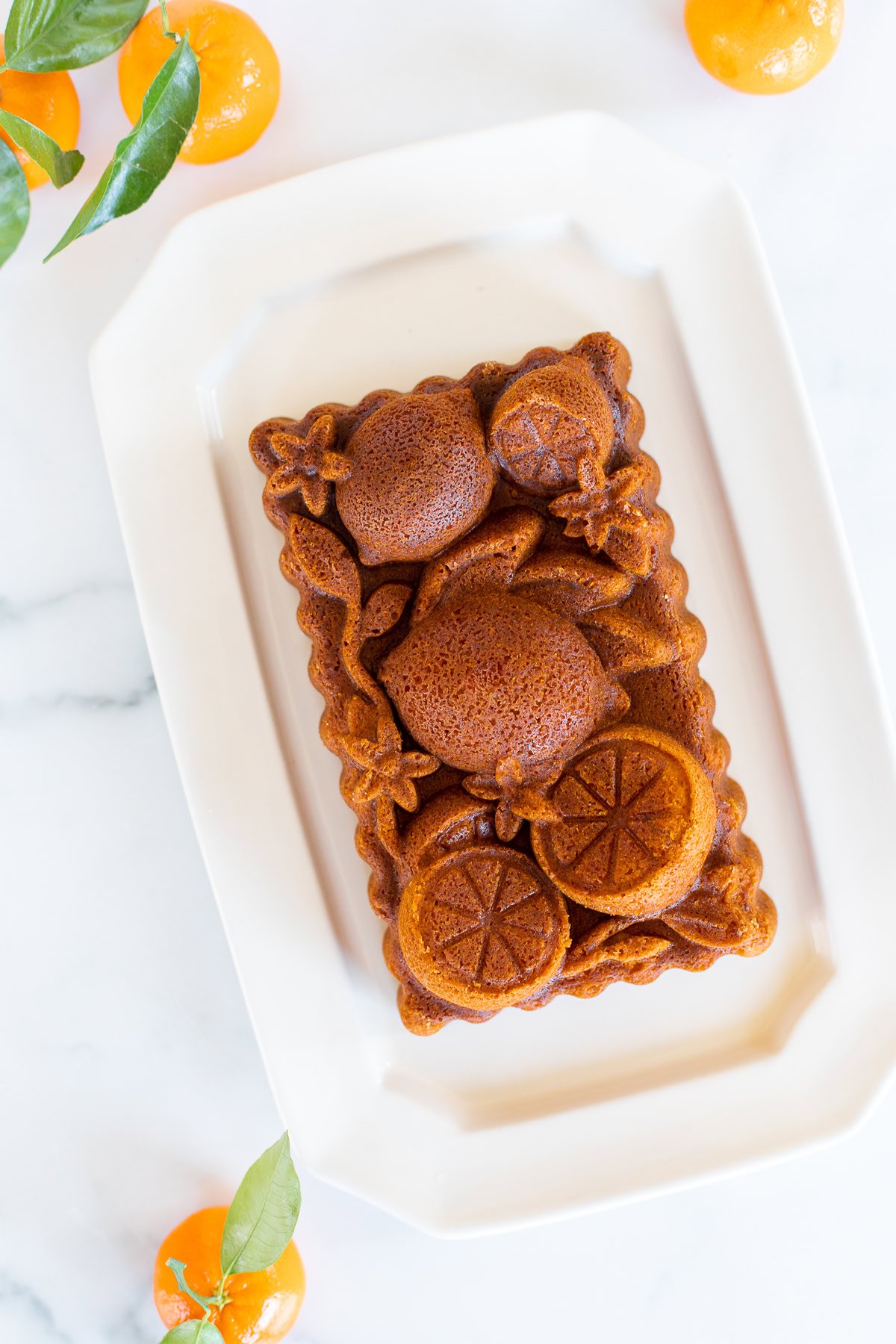 An orange pound cake on a white platter, surrounded by fresh oranges with green leaves. 