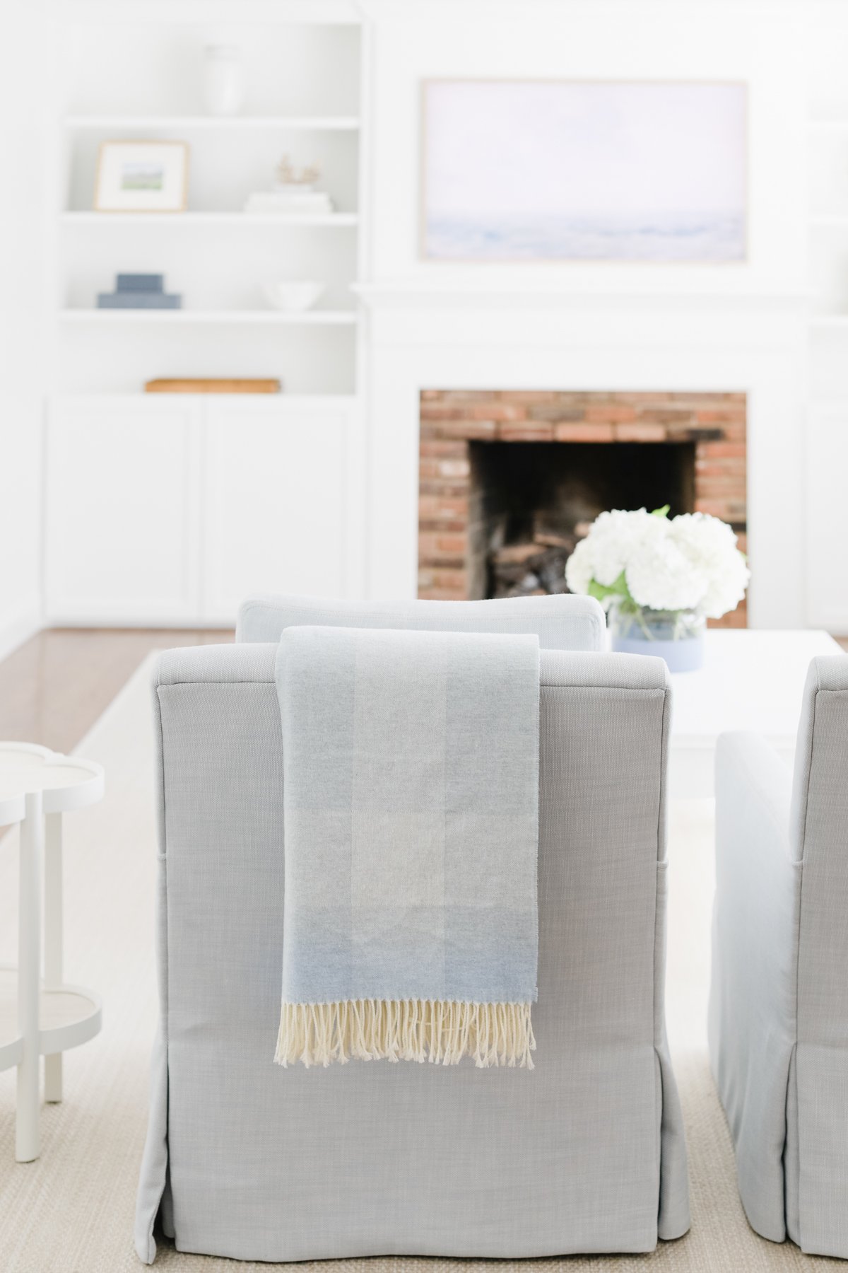 A living room with a brick fireplace, white shelves, cozy armchairs with a blanket draped over one, and a table with a vase of white flowers.