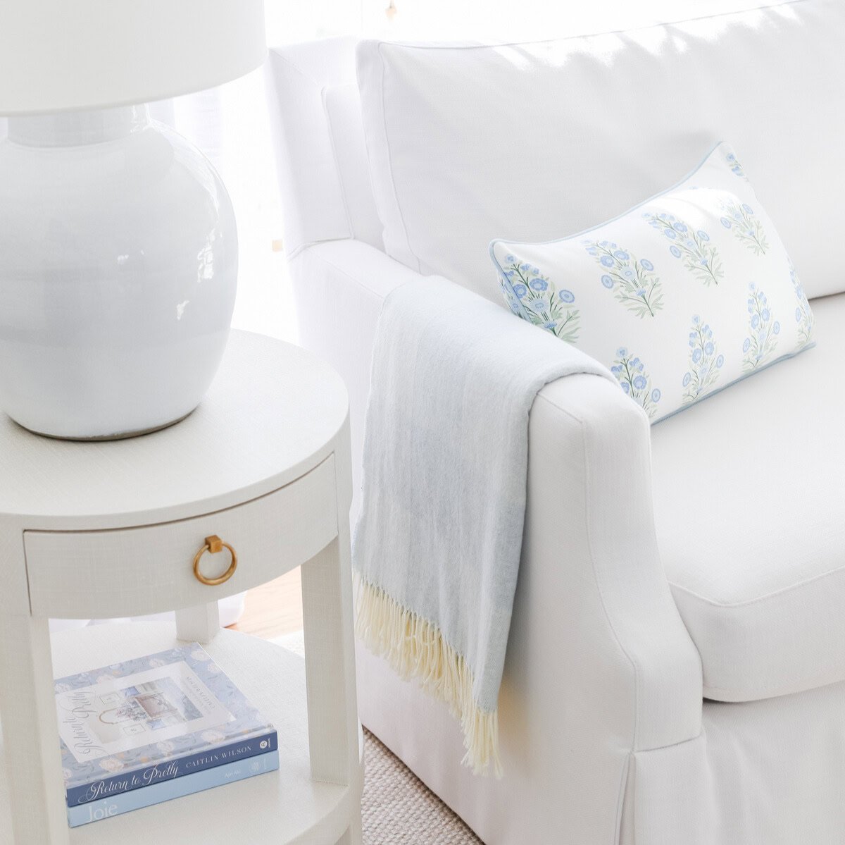 A white sofa with a blue sofa throw on the arm, next to a white side table with a large lamp. 