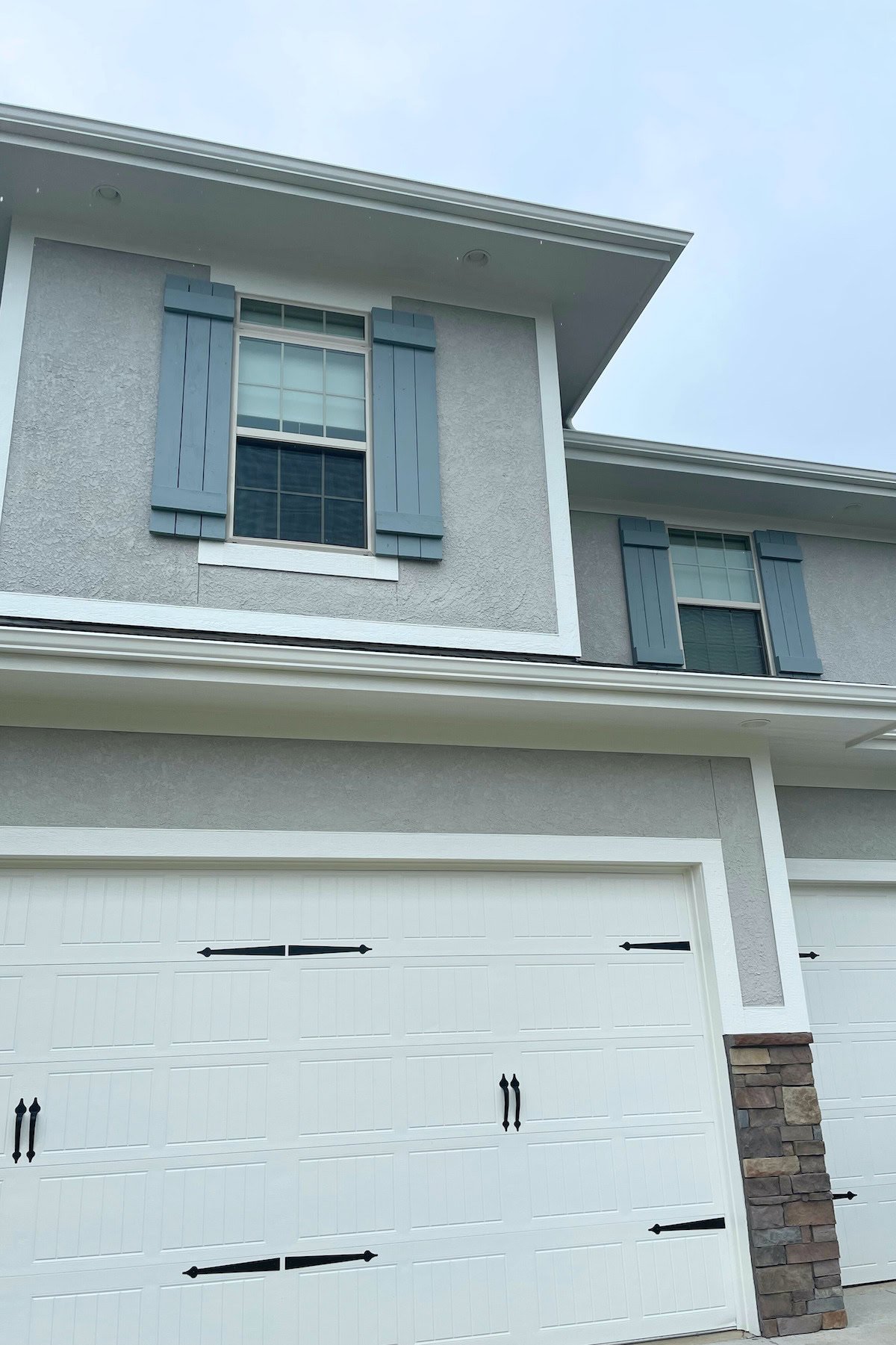 The two-story house features light blue shutters on the upper windows and a white garage door beneath. The exterior walls, painted in Benjamin Moore Brewster Gray, beautifully complement a section of brickwork to the side of the garage.