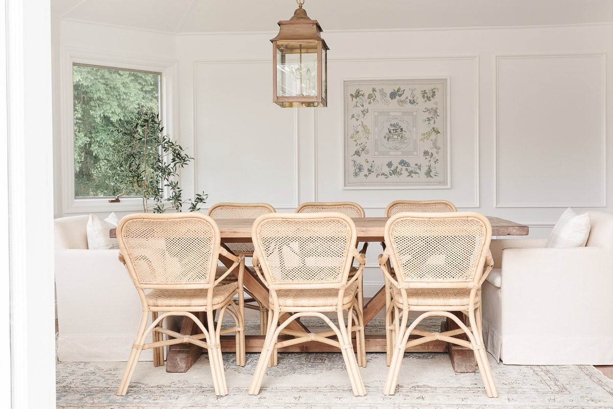 A dining room with light wooden chairs and a rectangular table, surrounded by beige upholstered seating. A hanging lantern-style light fixture and botanical Above it hangs a framed floral print on walls painted in a Benjamin Moore white color