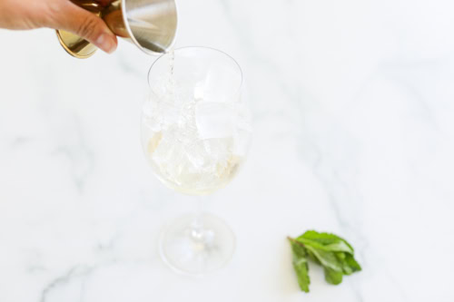 A hand pours liquid from a jigger into a wine glass filled with ice cubes. Fresh mint leaves are placed beside the glass on the white surface.