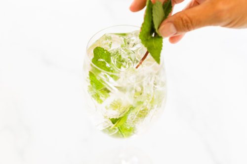 A hand placing mint leaves into a glass filled with ice and a Hugo rosé spritz.