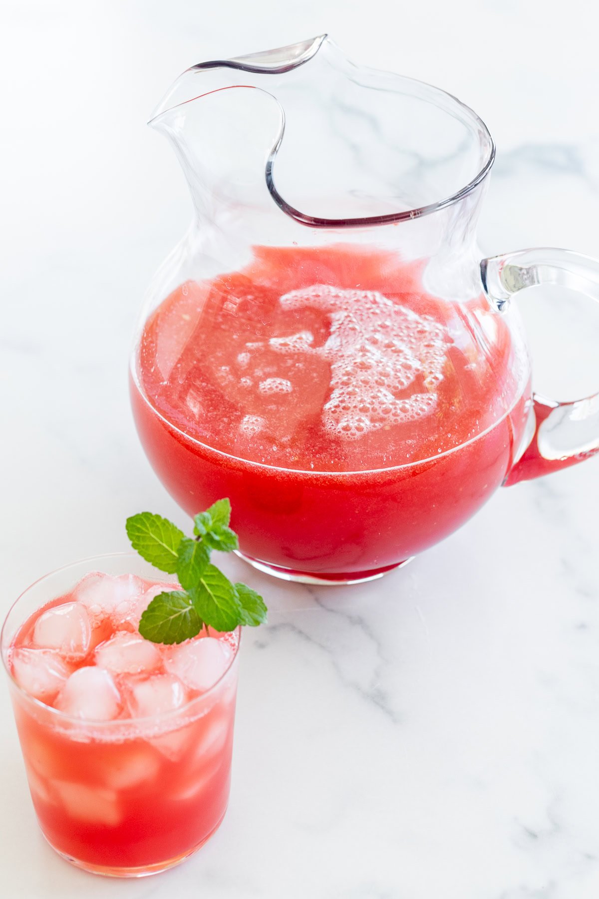 A glass pitcher filled with watermelon juice and a glass of the same juice with ice cubes and a mint garnish, both placed on a white surface.