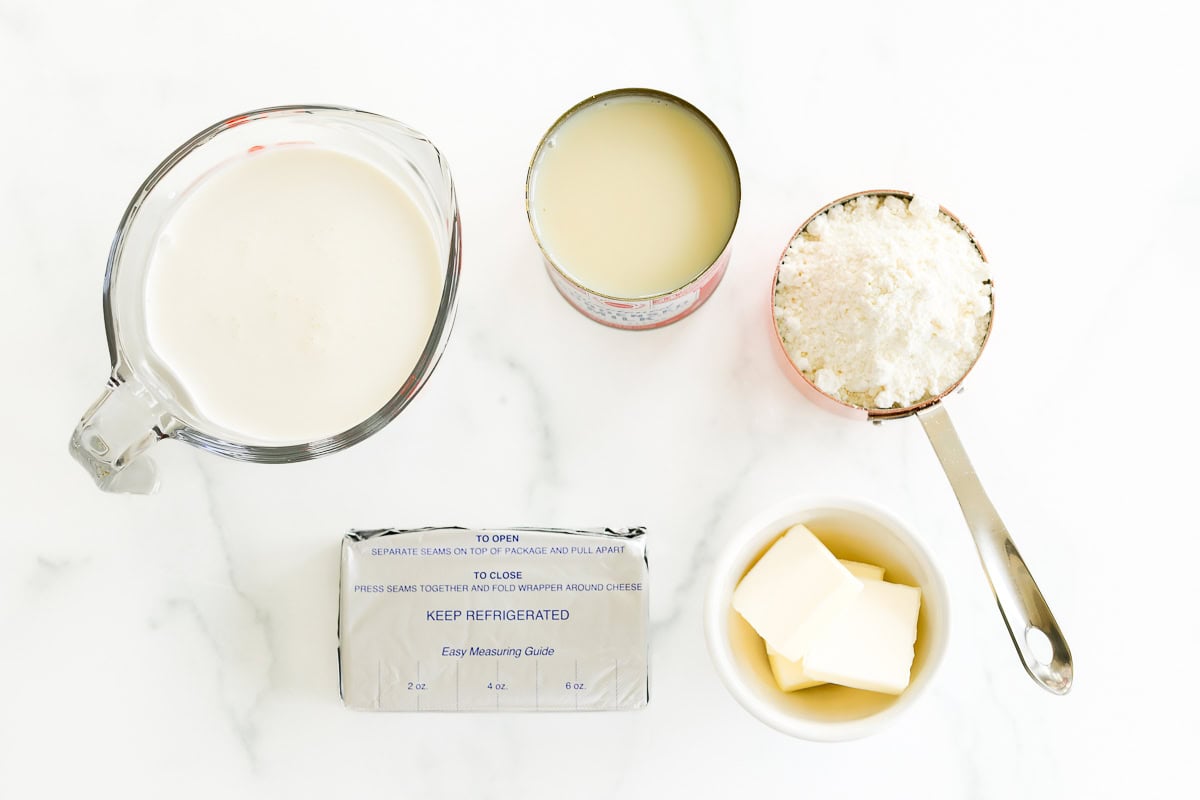 Ingredients for a Gooey Butter Cake Ice Cream recipe laid out on a white surface: a glass measuring cup with milk, an unopened can of condensed milk, a cup of grated coconut, a block of butter with wrapper, and butter pieces in a bowl.