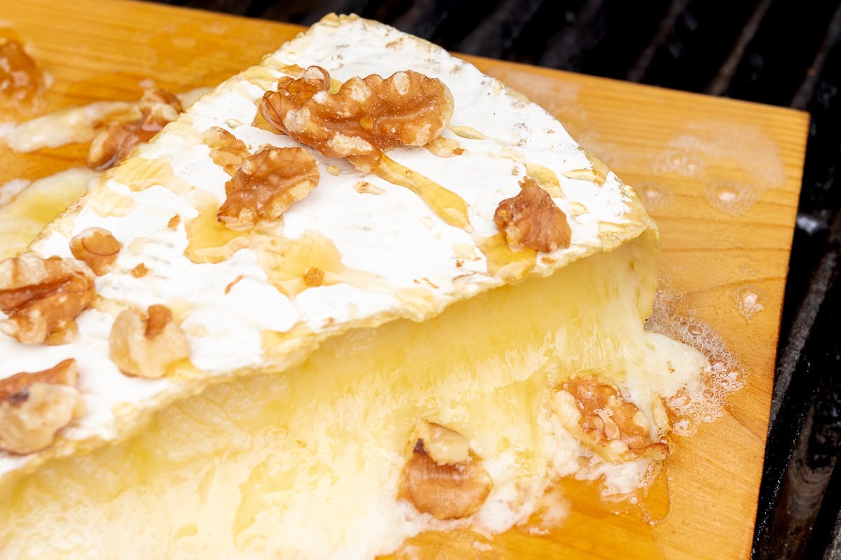 A wedge of grilled brie cheese topped with walnuts and honey on a wooden board, melting at the edges.