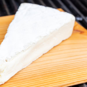 A wedge of soft cheese with a white rind sits on a wooden board.