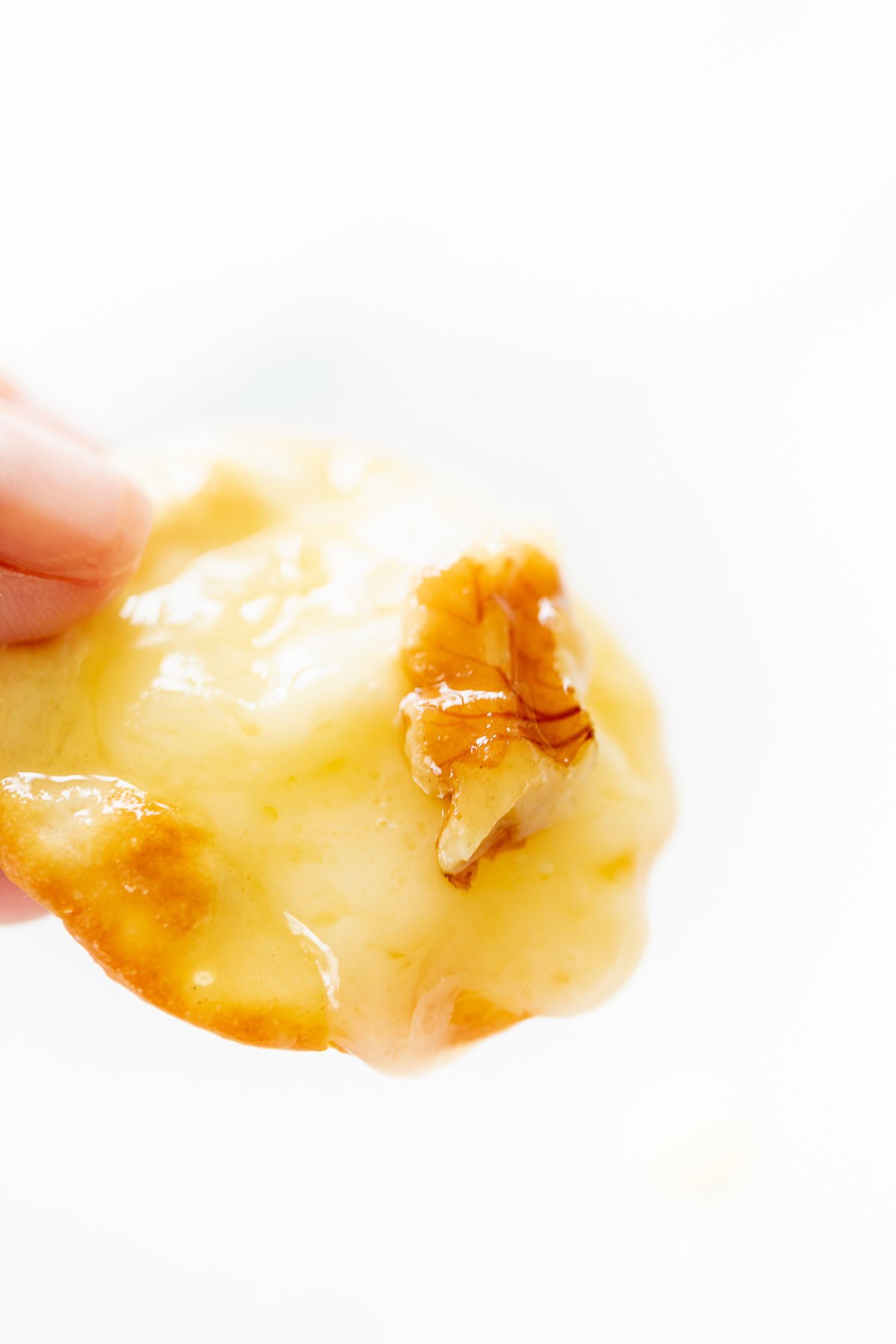 Close-up of a hand holding a cracker topped with melted grilled brie cheese and a walnut piece.