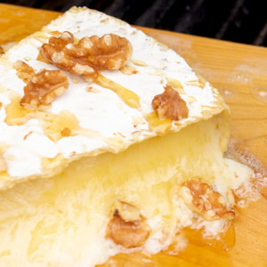 A wedge of melted brie cheese topped with honey and walnut pieces on a wooden board.