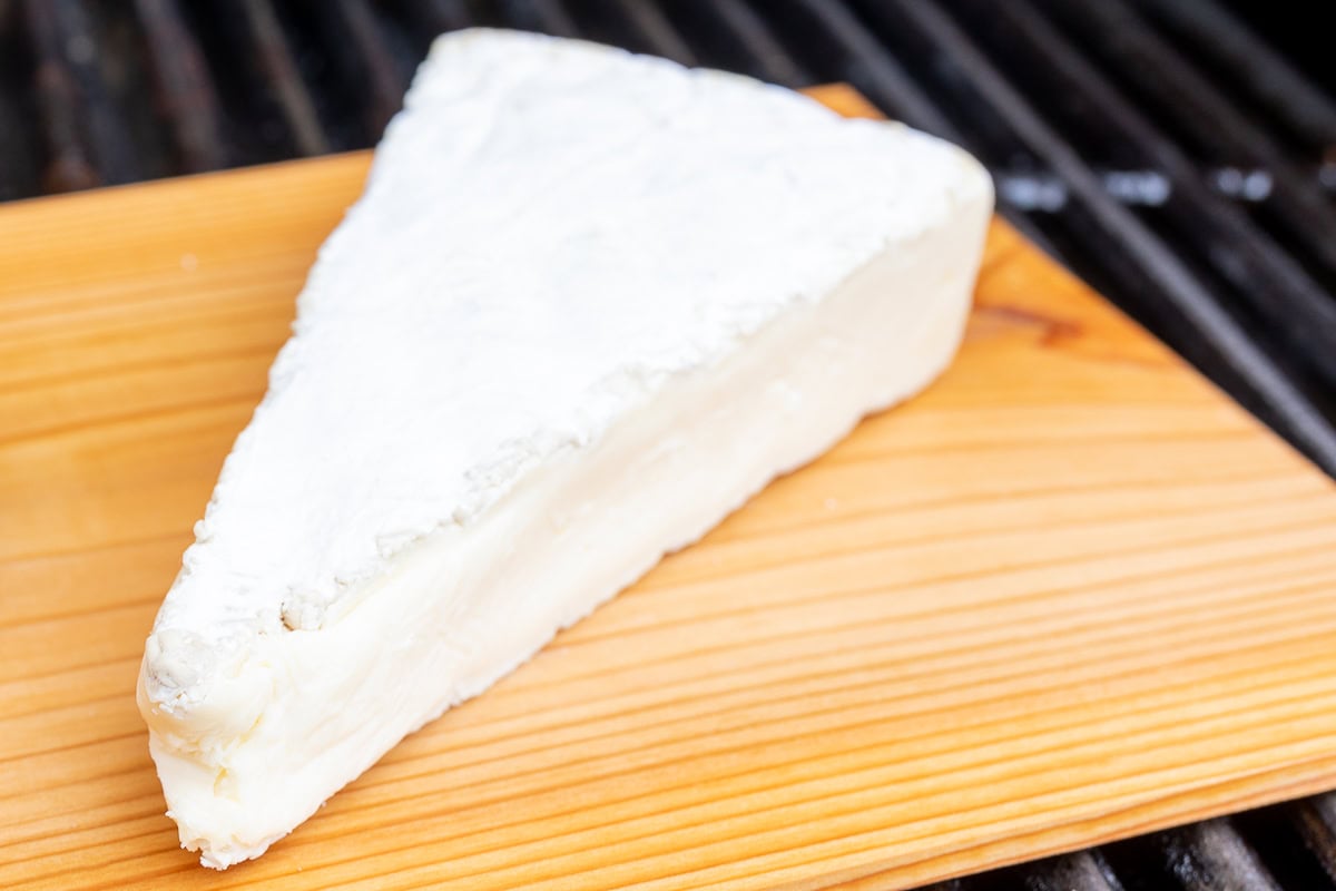 A wedge of soft cheese with a white rind sits on a wooden board.