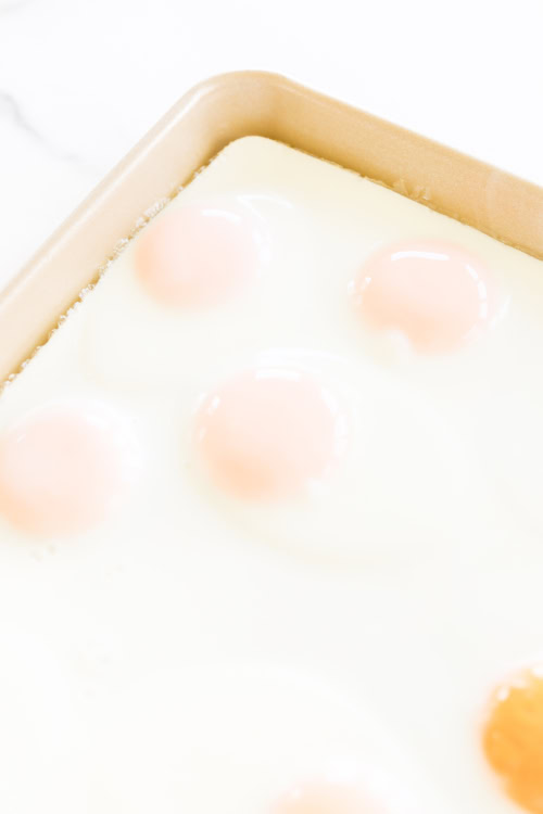 Close-up of several sunny side up eggs cooking on a light-colored skillet, resembling sheet pan eggs. The egg whites are set, and the yolks are soft and pinkish.