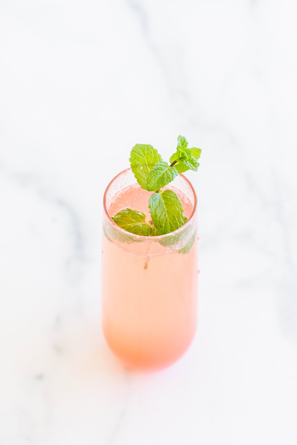 A tall glass of watermelon mimosa garnished with fresh mint leaves sits on a white marble surface.