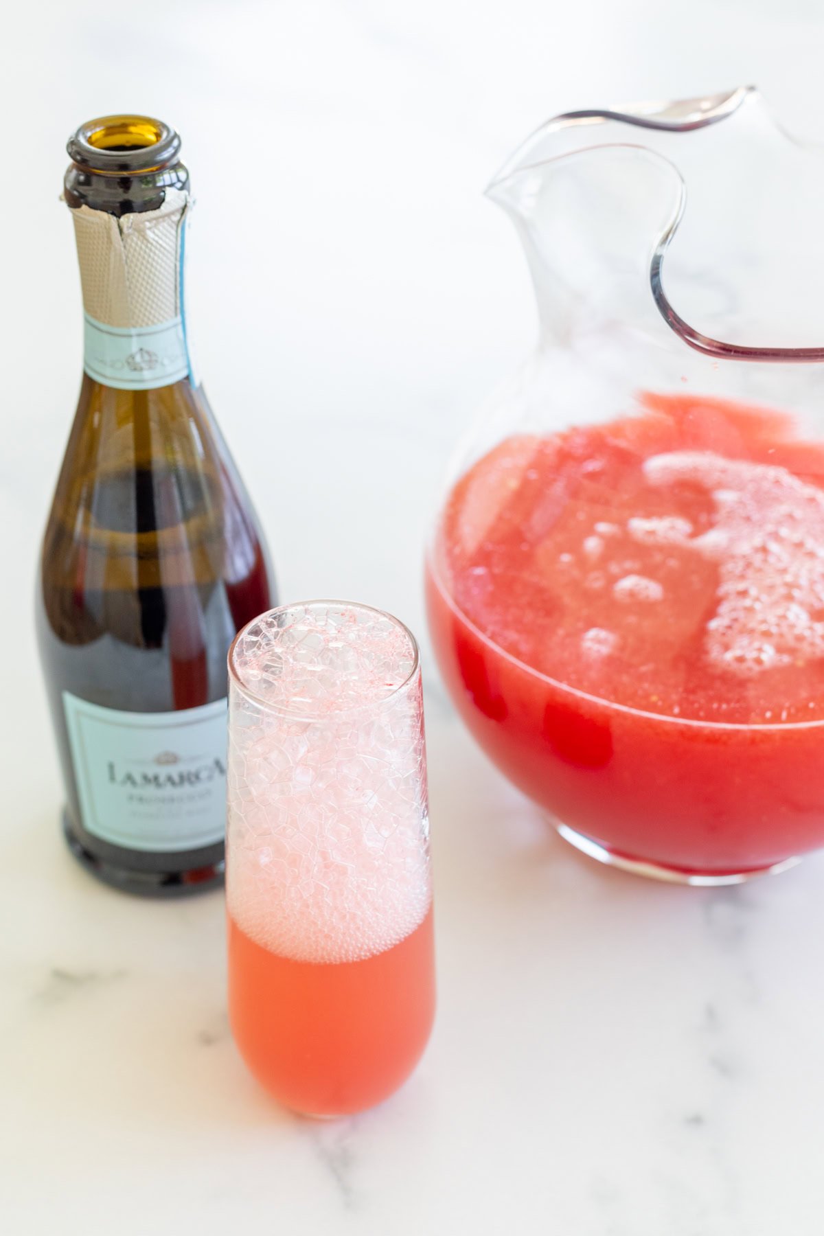 A bottle of La Marca prosecco, a glass of pink watermelon mimosa, and a pitcher filled with the same drink are arranged on a white countertop.