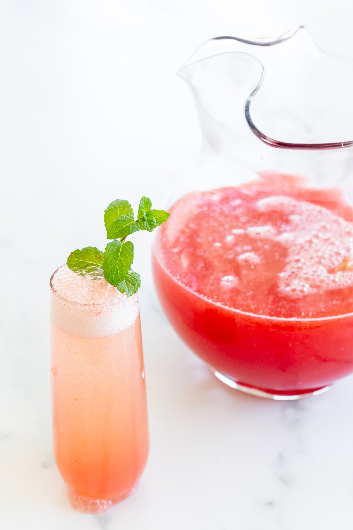 A glass of fizzy pink watermelon mimosa garnished with a mint sprig is placed next to a pitcher filled with the same refreshing drink.