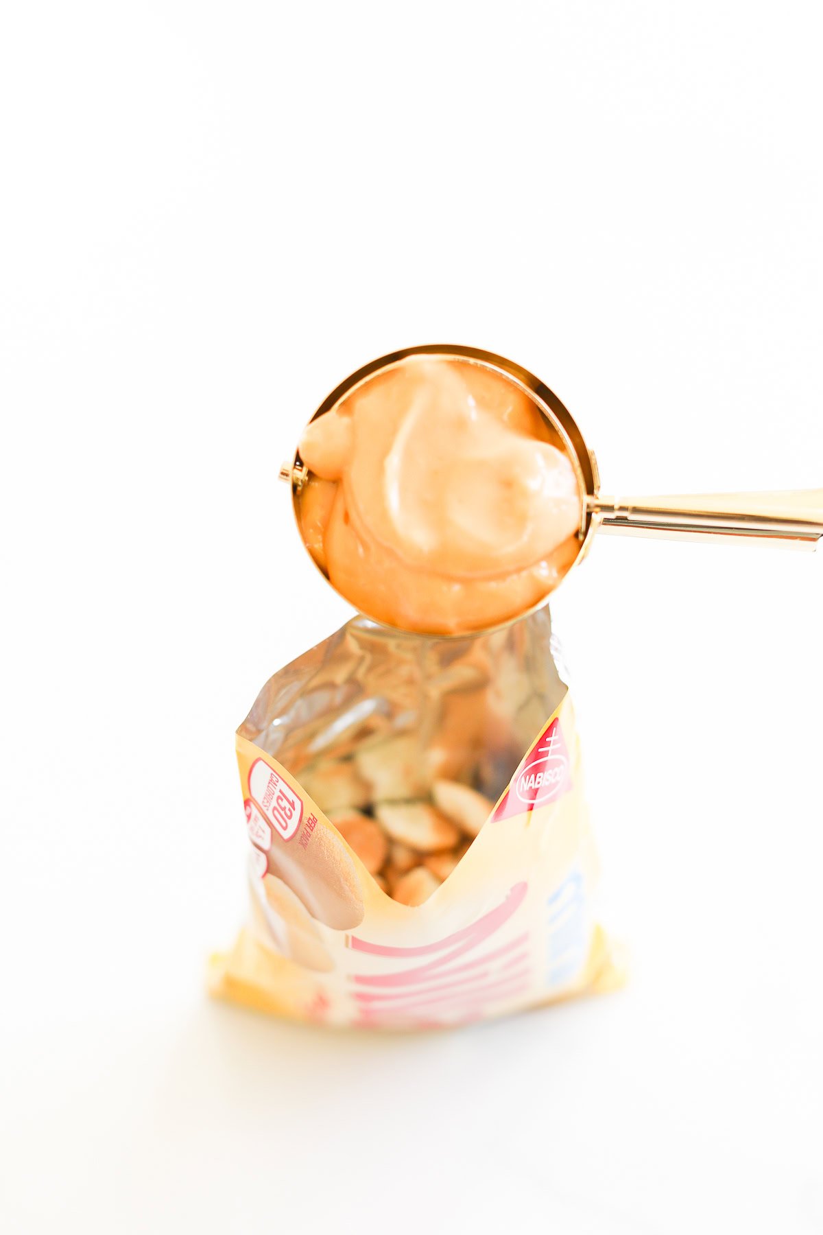 A cookie scoop filled with creamy peanut butter hovers over an opened bag of mini Nilla wafers, evoking the delightful essence of a playful butterscotch pie in a bag, all set against a pristine white background.