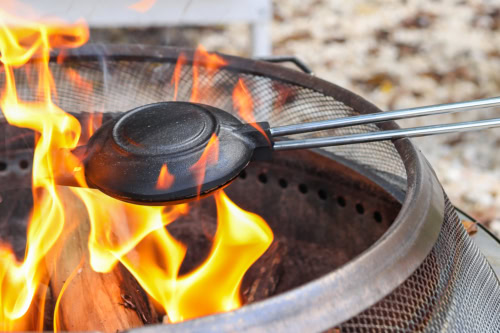 A metal pie iron, perfect for crafting a delicious pudgy pie, is held over an open flame in a fire pit, surrounded by burning wood.