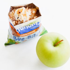 An open apple pie in a bag topped with whipped cream and nuts sits enticingly next to a crisp green apple on a white surface.