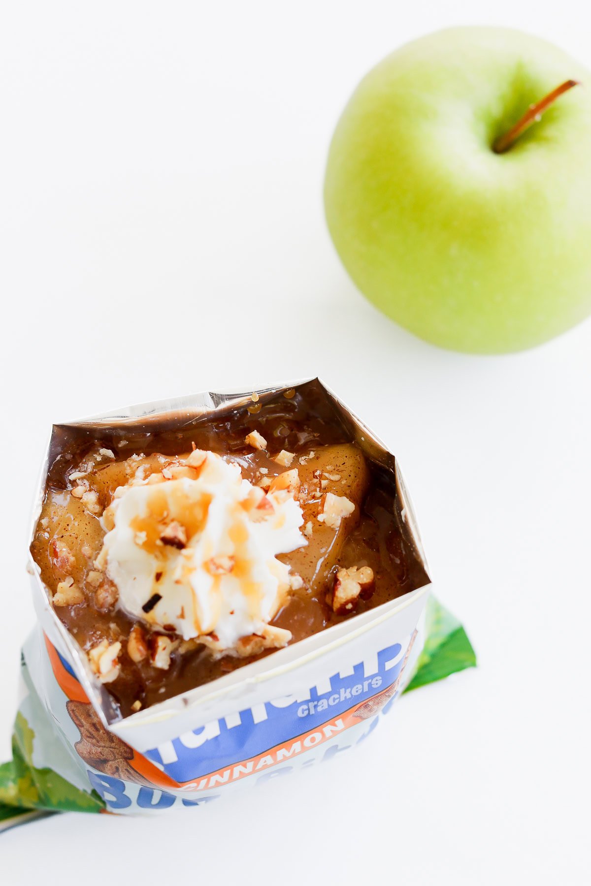 An open snack pack with whipped cream and nuts sits enticingly next to a crisp green apple on a white surface, reminiscent of an apple pie in a bag.