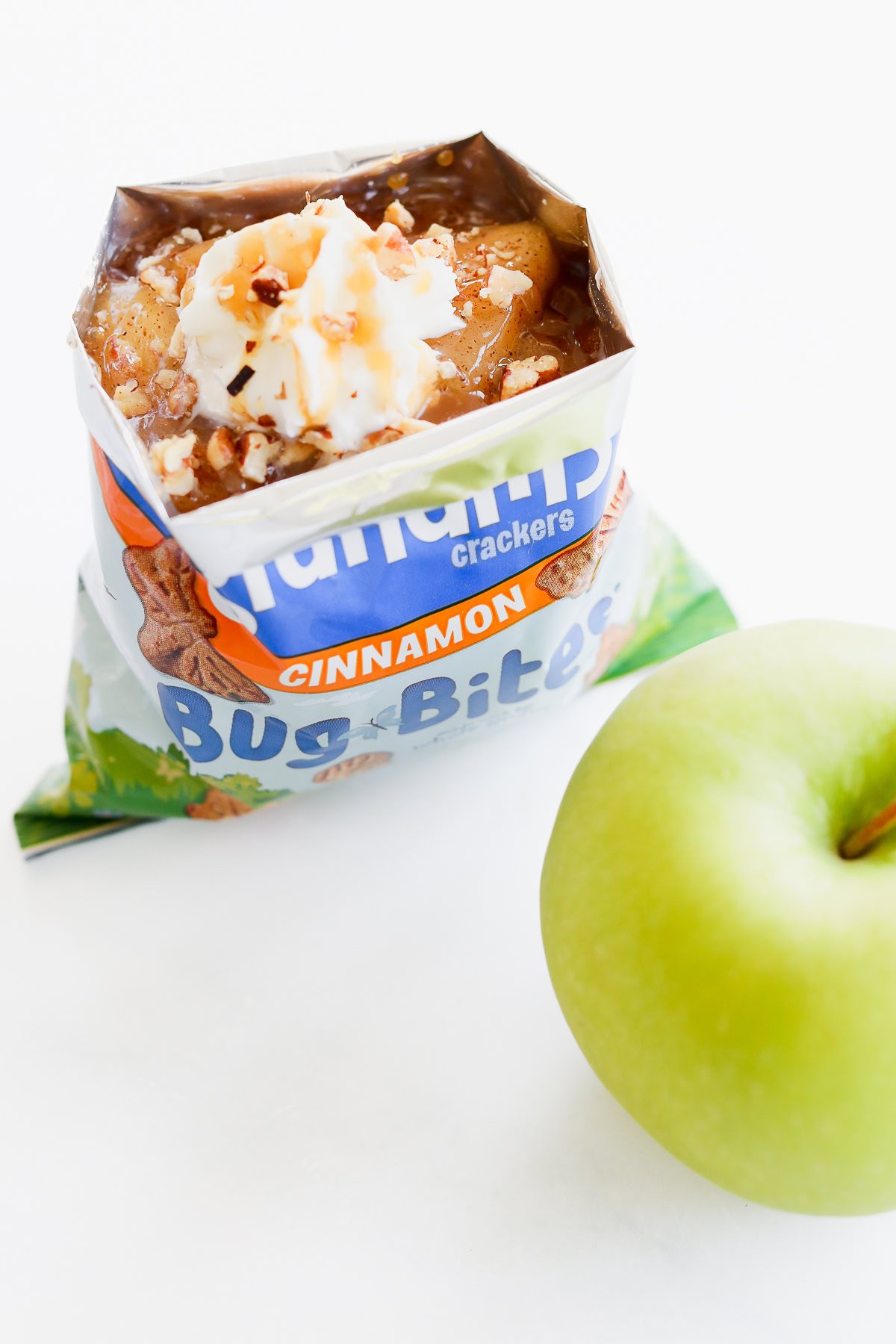 An open apple pie in a bag topped with whipped cream and nuts sits enticingly next to a crisp green apple on a white surface.