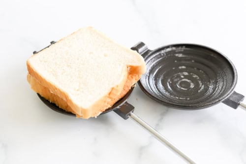 A slice of white bread nestles in an open round cast iron sandwich press on a white surface, ready to transform into a delicious pudgy pie.