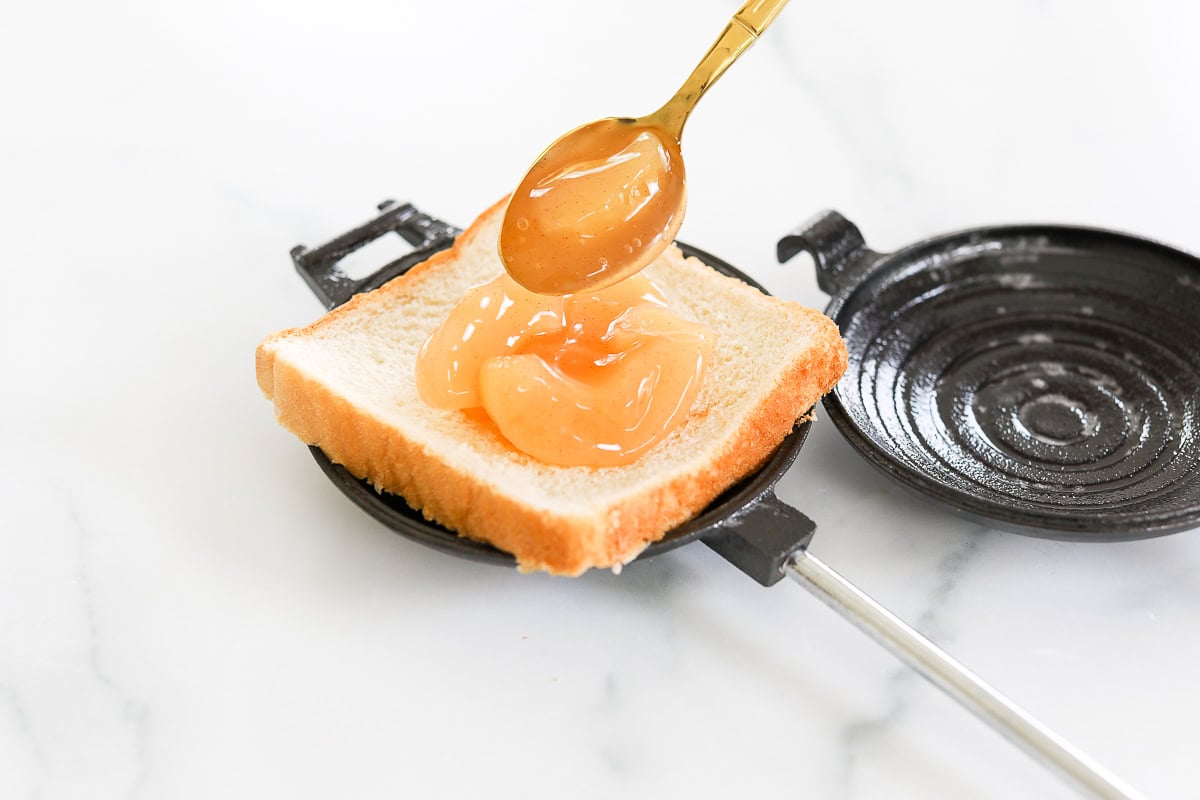 A spoonful of pie filling being spread on a slice of white bread placed in a black sandwich maker on a white surface.