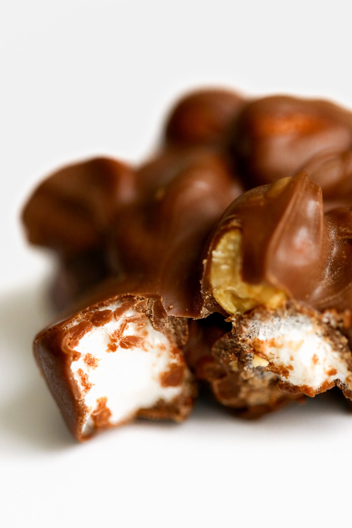 Close-up of a Rocky Road Candy chocolate cluster with a marshmallow filling, partially bitten to reveal the white interior, set against a plain background.