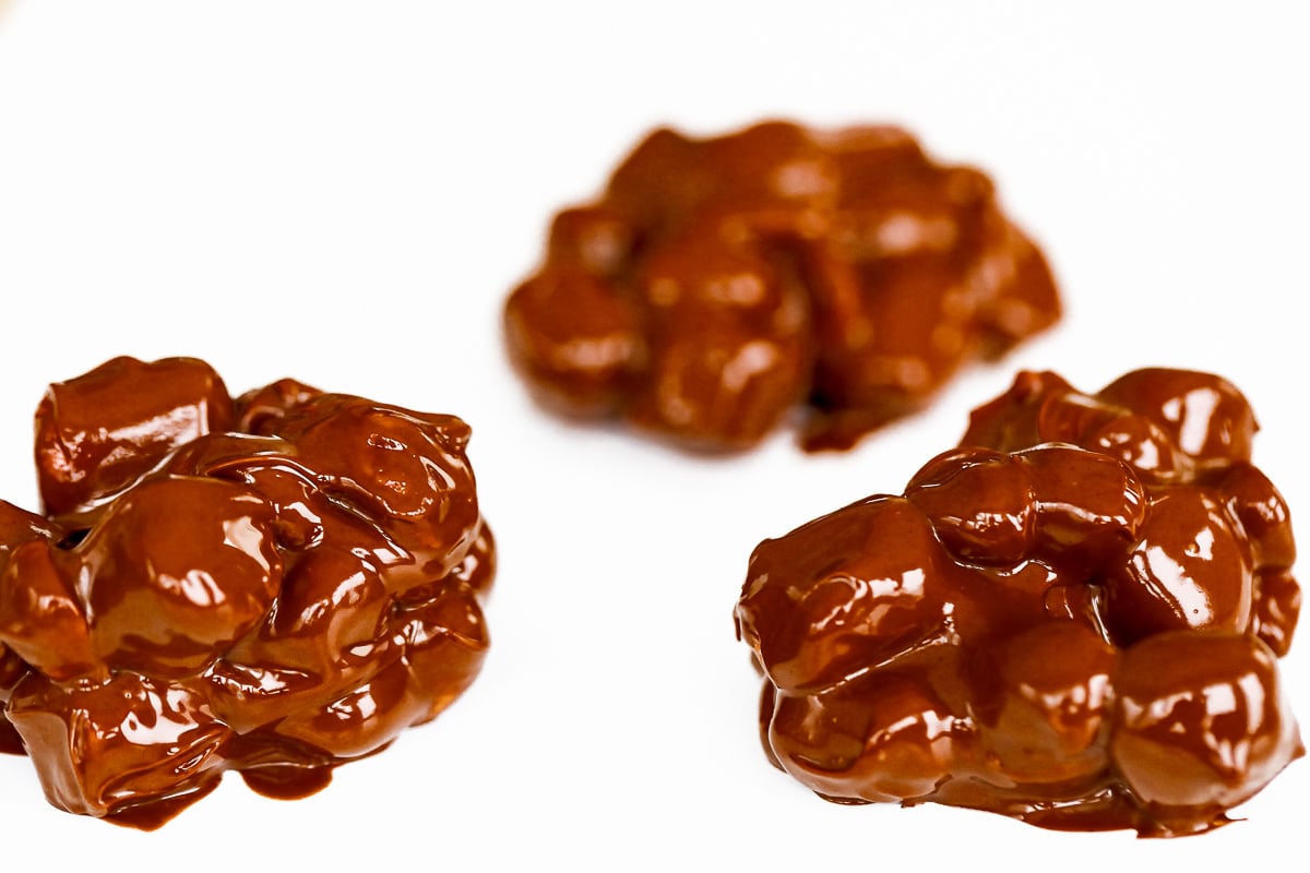 Three clusters of chocolate-covered nuts reminiscent of Rocky Road Candy rest on a white surface.