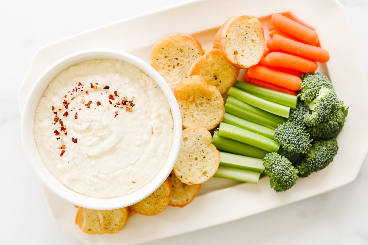 A platter featuring Asiago Cheese Dip paired with hummus, topped with red pepper flakes, and surrounded by sliced baguette, carrot sticks, celery sticks, and broccoli florets.